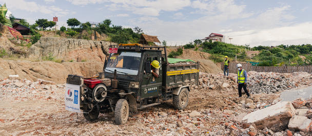 Small dump truck on a rocky construction site with a worker in a hard hat nearby.