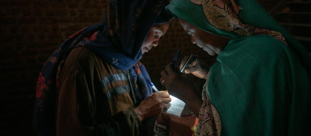 Photograph: two women in colorful headscarves lean toward a small light in a dim brick room.